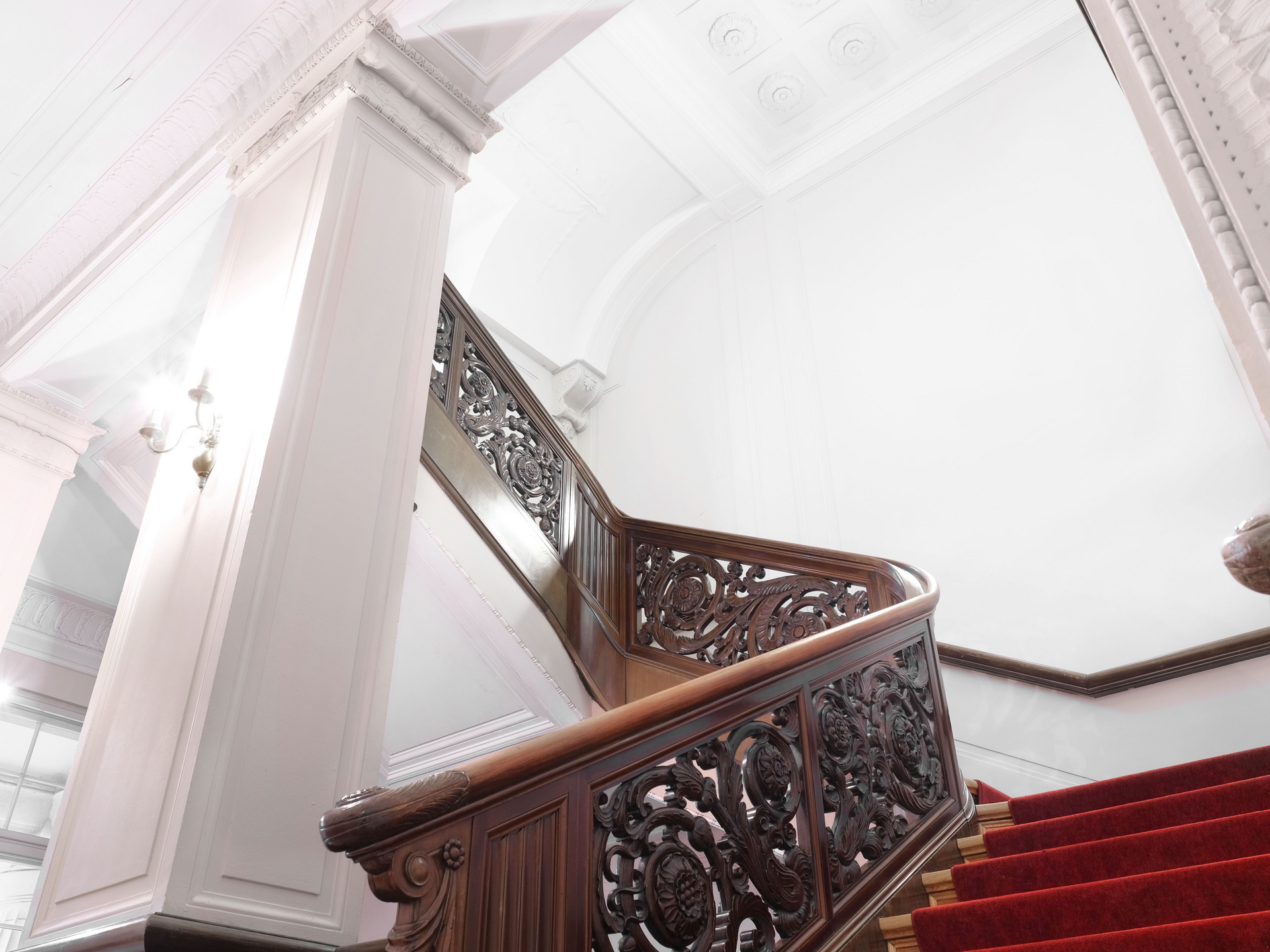 Ein prächtiges Treppengeländer aus dunkelbraunem Holz mit aufwendig geschnitzten Ornamenten führt eine Treppe mit rotem Teppich hinauf. Die helle weiße Wand und die verzierte Stuckdecke betonen den eleganten, historischen Charakter des Gebäudes.