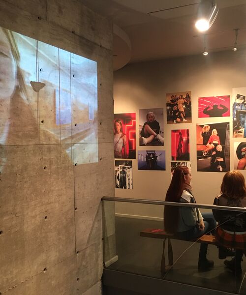 Eine Galerieausstellung mit Fotografien an der Wand. Links wird ein großes Bild auf eine Betonwand projiziert, während zwei Personen auf einer Bank sitzen und die Ausstellung betrachten.