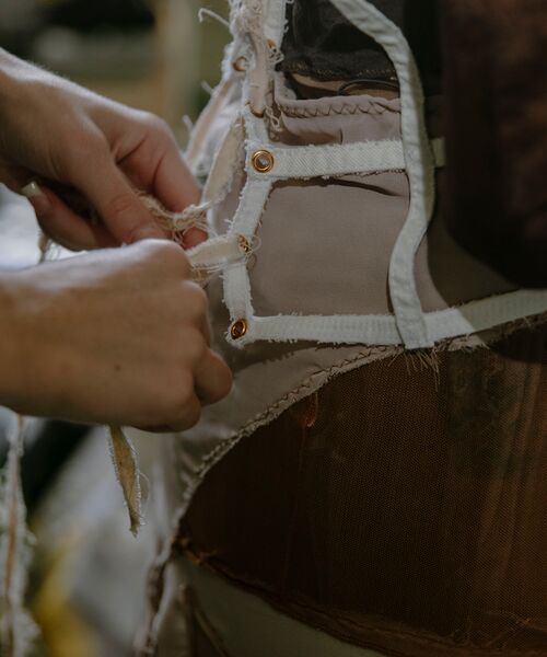 Nahaufnahme von Händen, die an einem Kleidungsstück arbeiten – Stoff wird festgehalten oder genäht, sichtbare Nähte, Knopflöcher und verschiedene Materialien.