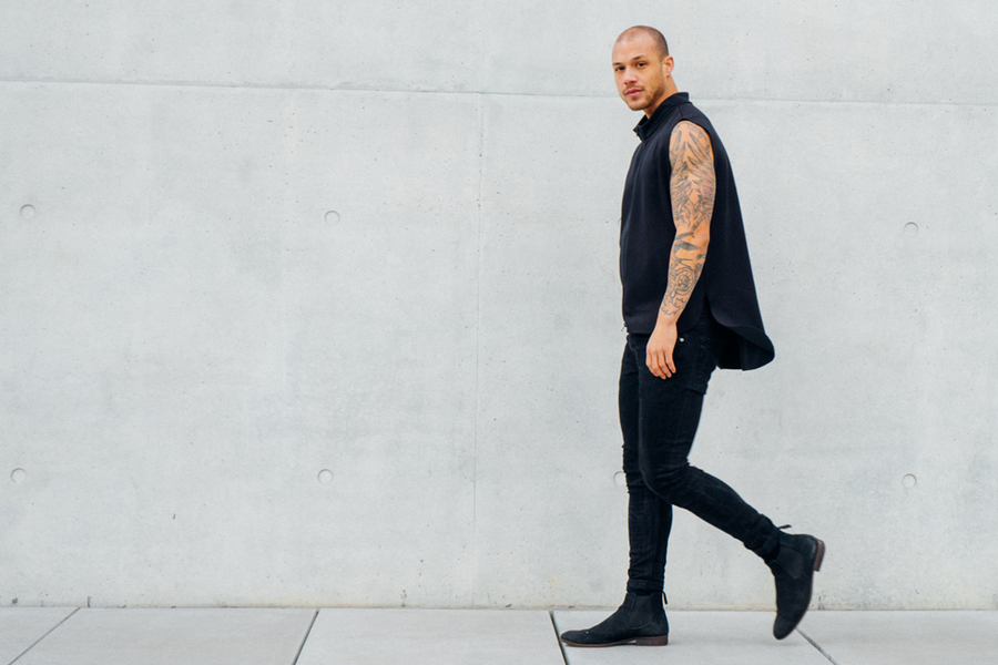 Mann mit tätowiertem Arm in schwarzer Kleidung geht vor einer Betonwand.