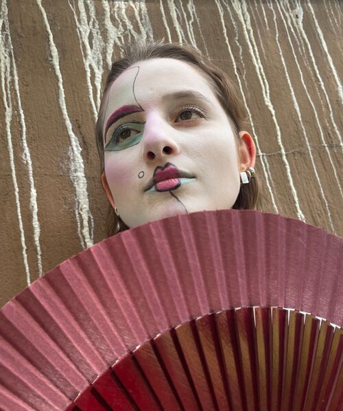 Porträt einer Person mit künstlerischem Gesichtsmakeup und einem roten Fächer im Vordergrund.