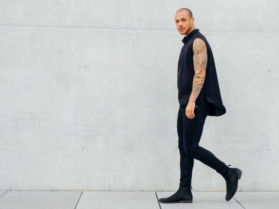 Mann mit tätowiertem Arm in schwarzer Kleidung geht vor einer Betonwand.