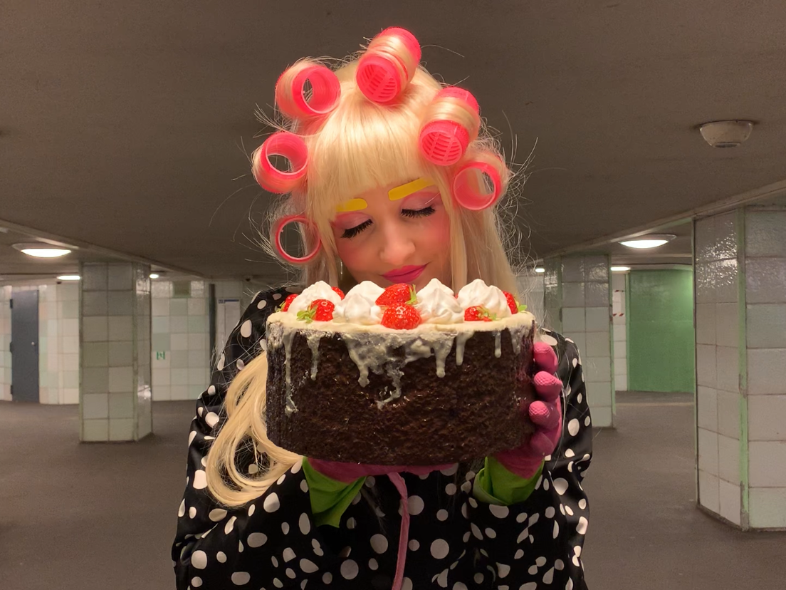 Das Bild zeigt eine Person in einem U-Bahn-ähnlichen, gekachelten Untergrundgang. Sie trägt blonde Perücke mit großen rosa Lockenwicklern, auffälliges Make-up mit bunten Augenbrauen und Wimpern sowie pinke Handschuhe. Sie hält eine große, mit Sahne und Er