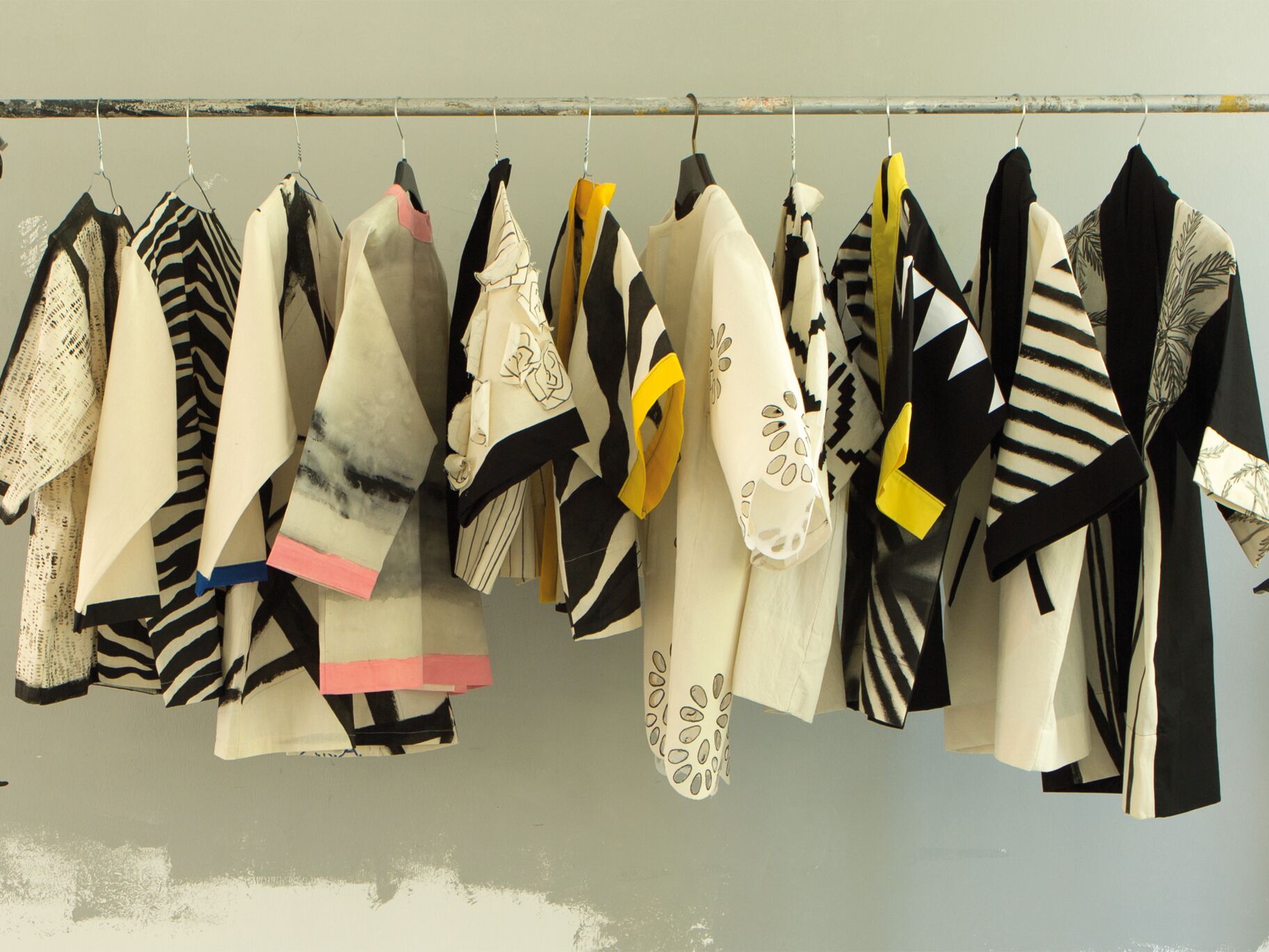 Mehrere kunstvoll gestaltete Kimono mit grafischen Mustern in Schwarz, Weiß und Beige hängen nebeneinander auf einer schlichten Kleiderstange vor grauer Wand.