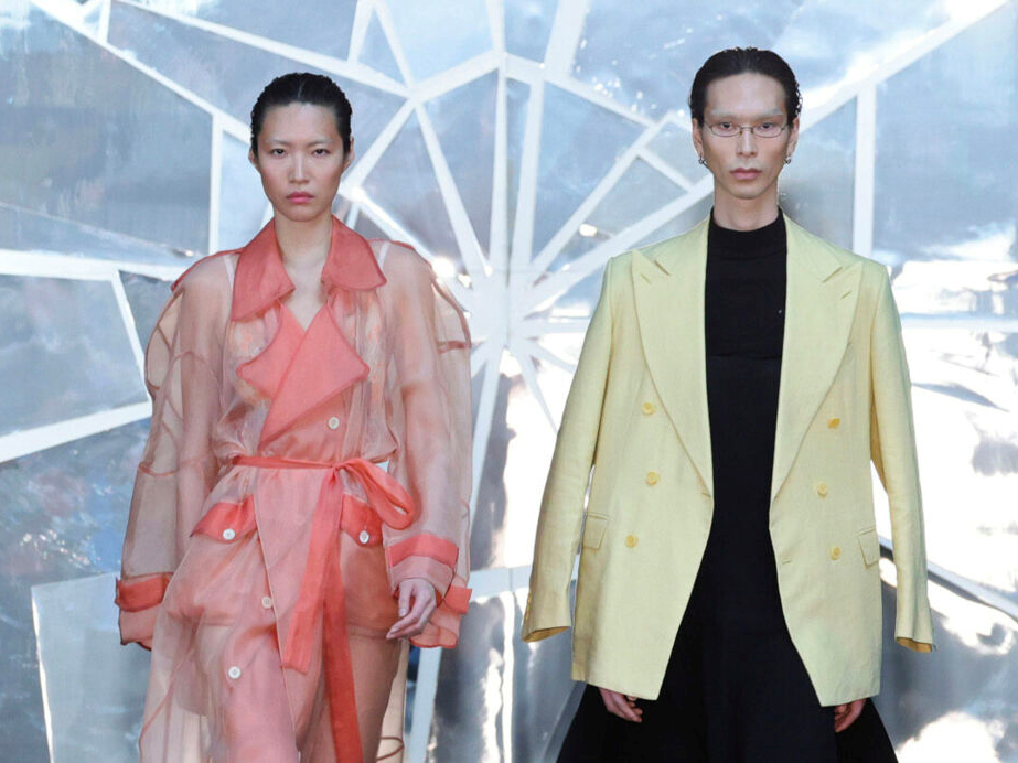 Zwei Models auf einem Laufsteg: Links trägt eine Person einen transparenten, lachsfarbenen Trenchcoat mit Bindegürtel, rechts eine Person einen hellgelben Blazer über schwarzem Outfit. Im Hintergrund eine geometrische silbrig-weiße Struktur.