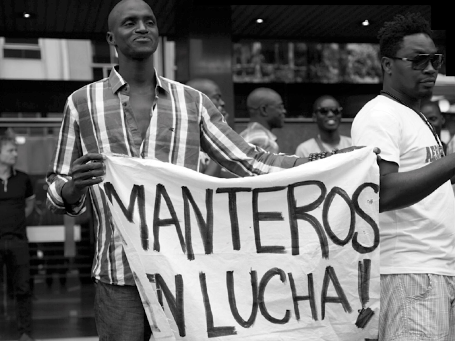 Schwarz-weiß-Foto einer Demonstration, bei der zwei Männer ein Transparent mit der Aufschrift „Manteros en lucha!“ hochhalten. Im Hintergrund sind weitere Personen zu sehen, einige mit Sonnenbrillen, die die Szene beobachten oder daran teilnehmen.