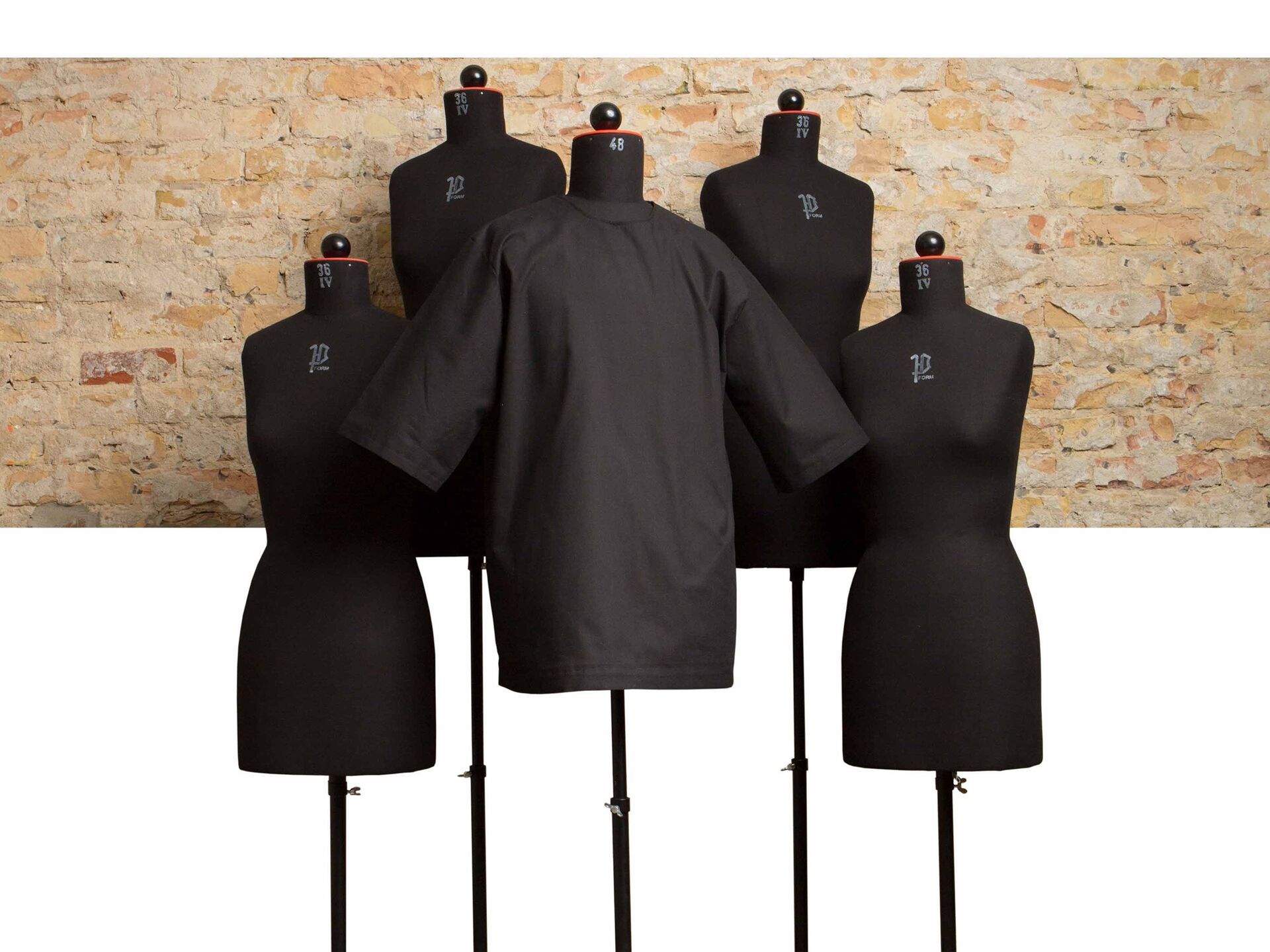 Fünf Schneiderpuppen vor einer Backsteinwand, eine davon trägt ein schwarzes T-Shirt.