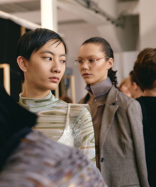 Zwei Personen in moderner, minimalistischer Mode stehen nebeneinander in einem hellen Raum, die vordere trägt ein gemustertes Oberteil, die hintere einen grauen Blazer mit Brille.