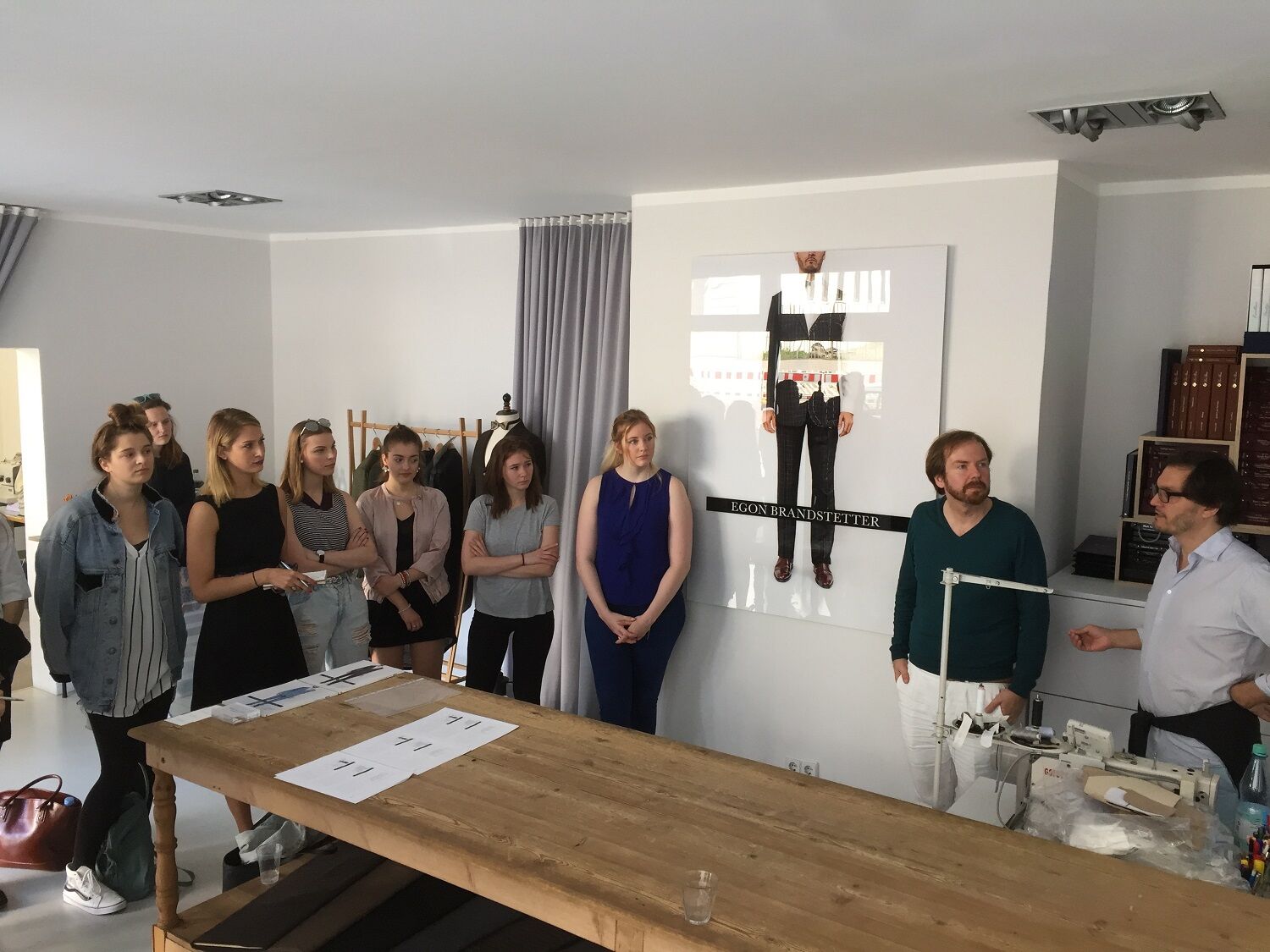 Dozent präsentiert Maßanzug-Anpassungen vor einer Gruppe, Projektion mit Anzugfotos im Hintergrund.