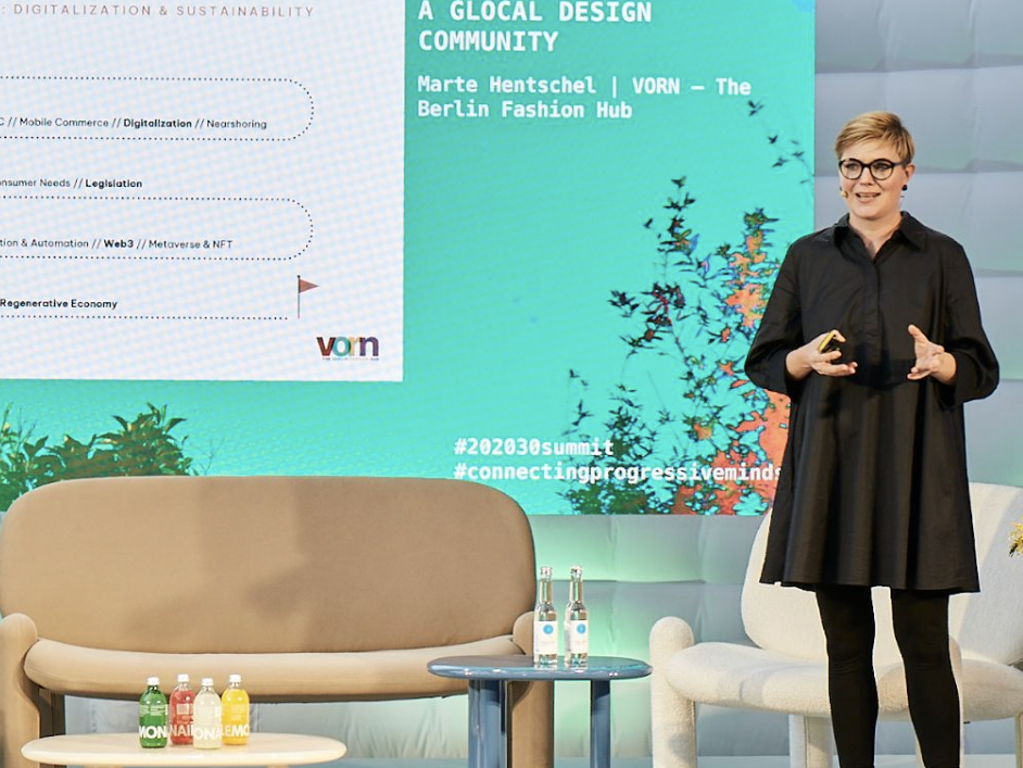 Eine Person in schwarzem Outfit steht auf einer Bühne und hält eine Präsentation. Hinter ihr ist eine große Leinwand mit Texten wie „A Glocal Design Community“, „VORN – The Berlin Fashion Hub“ sowie Schlagworten wie „Digitalization“, „Legislation“ und „We