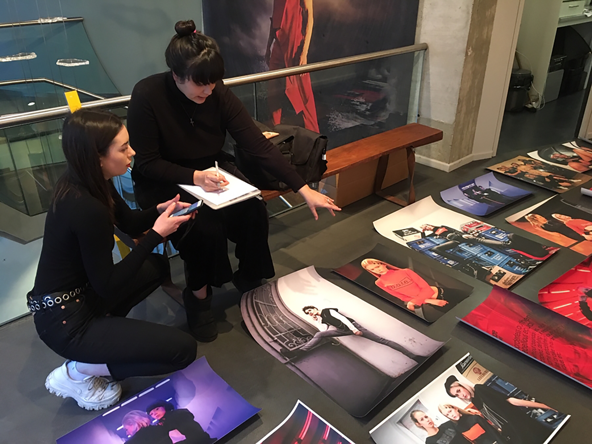 Zwei Frauen sitzen auf dem Boden einer Galerie und betrachten großformatige Fotografien, die vor ihnen ausgelegt sind. Eine Frau hält ein Handy, die andere macht Notizen in ein Heft und zeigt auf ein Bild. Es wirkt wie die Vorbereitung oder Planung einer