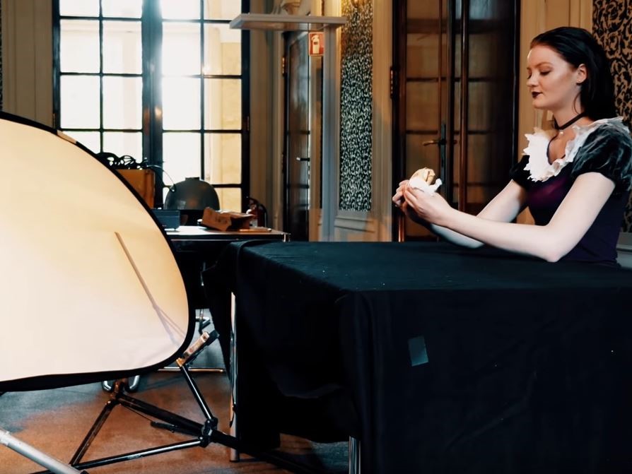 Eine Frau in einem Kostüm sitzt an einem schwarzen, mit Stoff bedeckten Tisch und hält einen kleinen Gegenstand in den Händen. Links im Bild ist ein großes Softbox-Licht für eine Fotosession oder Videodreh aufgestellt. Im Hintergrund sind große Fenster un