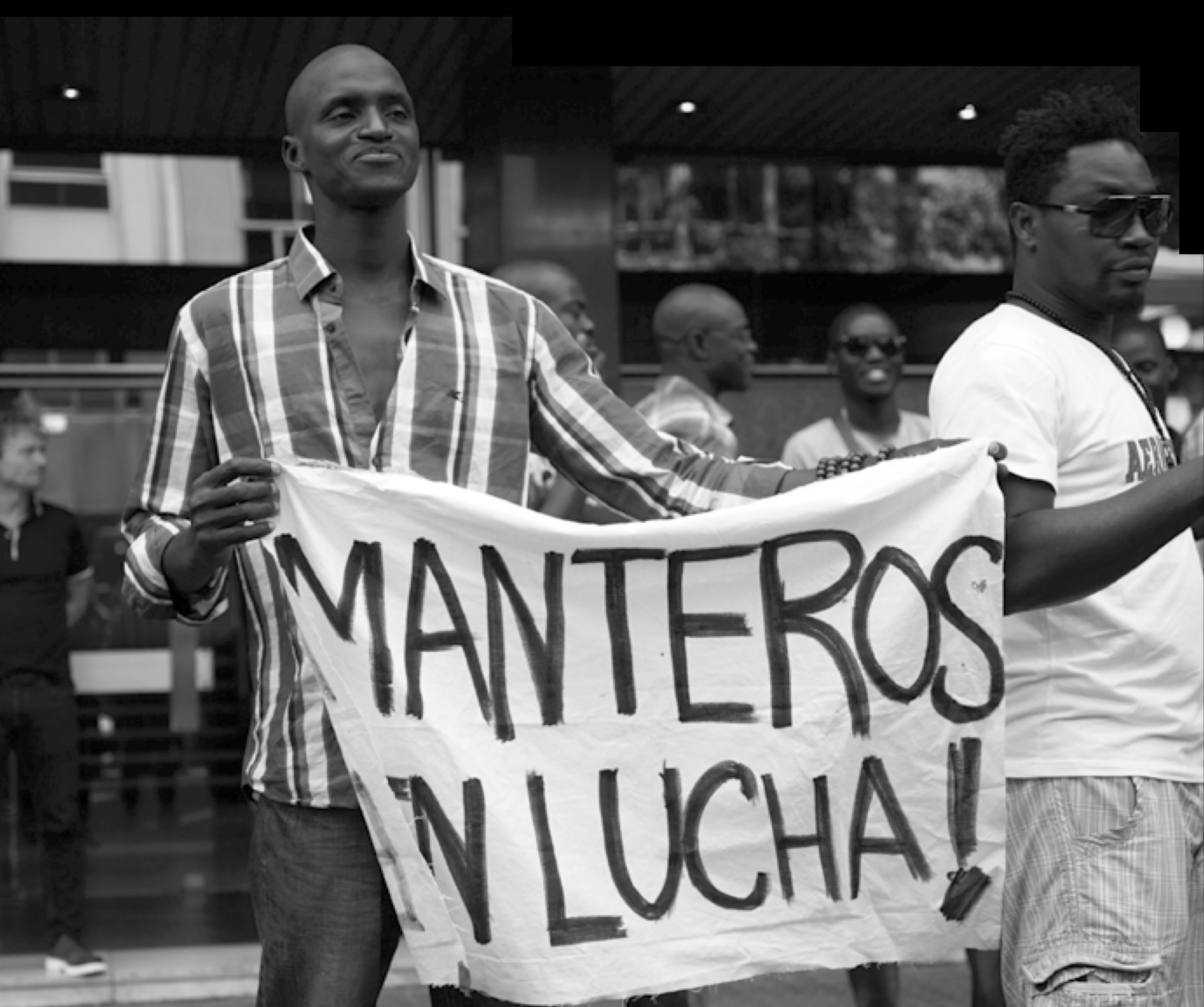 Schwarz-weiß-Foto einer Demonstration, bei der zwei Männer ein Transparent mit der Aufschrift „Manteros en lucha!“ hochhalten. Im Hintergrund sind weitere Personen zu sehen, einige mit Sonnenbrillen, die die Szene beobachten oder daran teilnehmen.
