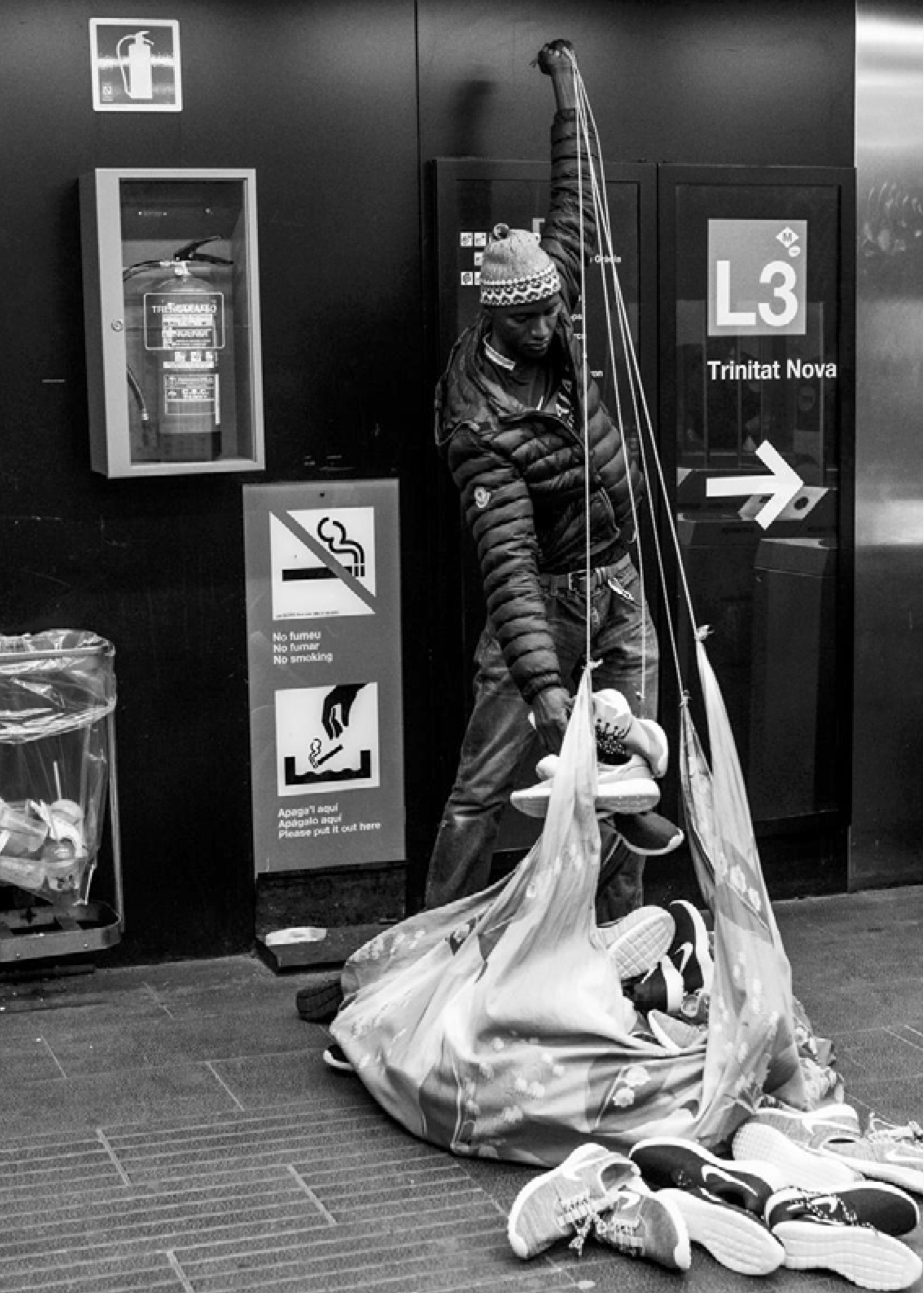 Schwarz-weiß-Foto eines Straßenverkäufers in einer U-Bahn-Station, der gefälschte Turnschuhe auf einem Tuch präsentiert. Im Hintergrund sind Hinweisschilder und ein Feuerlöscherkasten zu sehen, daneben ein Schild mit der Aufschrift „L3 Trinitat Nova“.